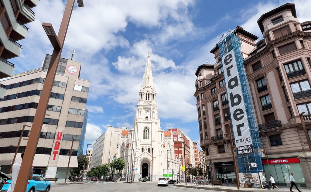 ビルバオ Bilbao 中央はサン・ホセ教会（Iglesia de San José Montana）。白い外観と鋭い尖塔が印象的なネオ・ゴシック様式の教会。周囲の近代的なオフィスビルや、ビルバオの労働組合「UGT」の看板との対比が、歴史と現代が共存するこの街を象徴している。