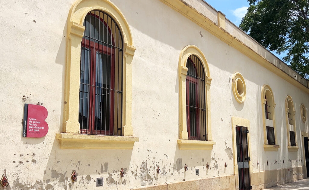 バルセロナ（Barcelona）バイ・グイナルド＝カン・バロ社会サービスセンター（Centre de Serveis Socials Baix Guinardó-Can Baró）施設の名称が記された赤い看板と、クリーム色の外壁に規則的に並ぶアーチ型の窓